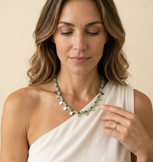 A woman wearing a chunky statement necklace made of mint green beads and small metallic gold and silver rectangular charms, styled with a white one-shoulder top against a warm beige background.