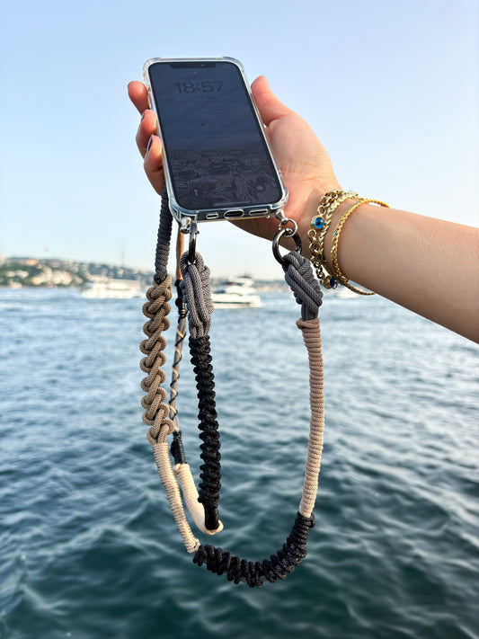 Hand holding a phone with a granite shore macrame crossbody strap against a scenic background of water and sky.