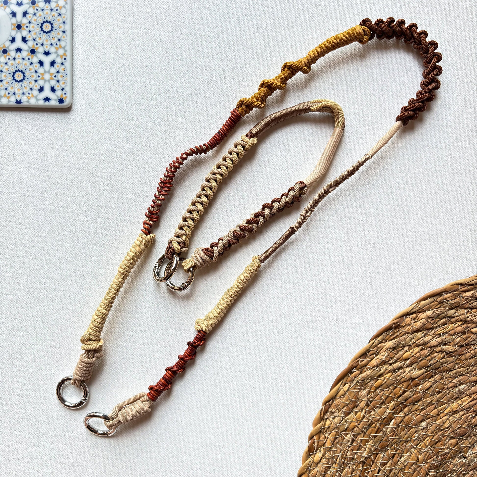 Three braided crossbody straps in beige, brown, and red on a white surface with a woven mat