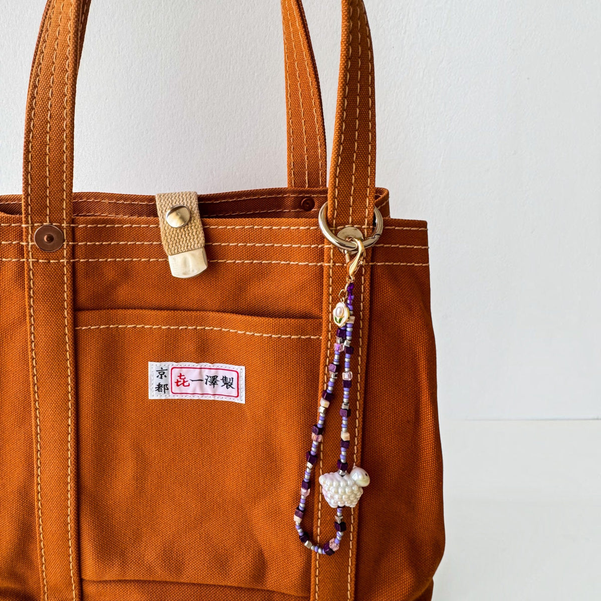 A close-up of an orange-brown canvas tote bag with white contrast stitching, a front pocket, a brass snap closure, and a decorative beaded keychain hanging from the side ring. The bag has a small Japanese label on the pocket.