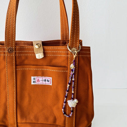 A close-up of an orange-brown canvas tote bag with white contrast stitching, a front pocket, a brass snap closure, and a decorative beaded keychain hanging from the side ring. The bag has a small Japanese label on the pocket.