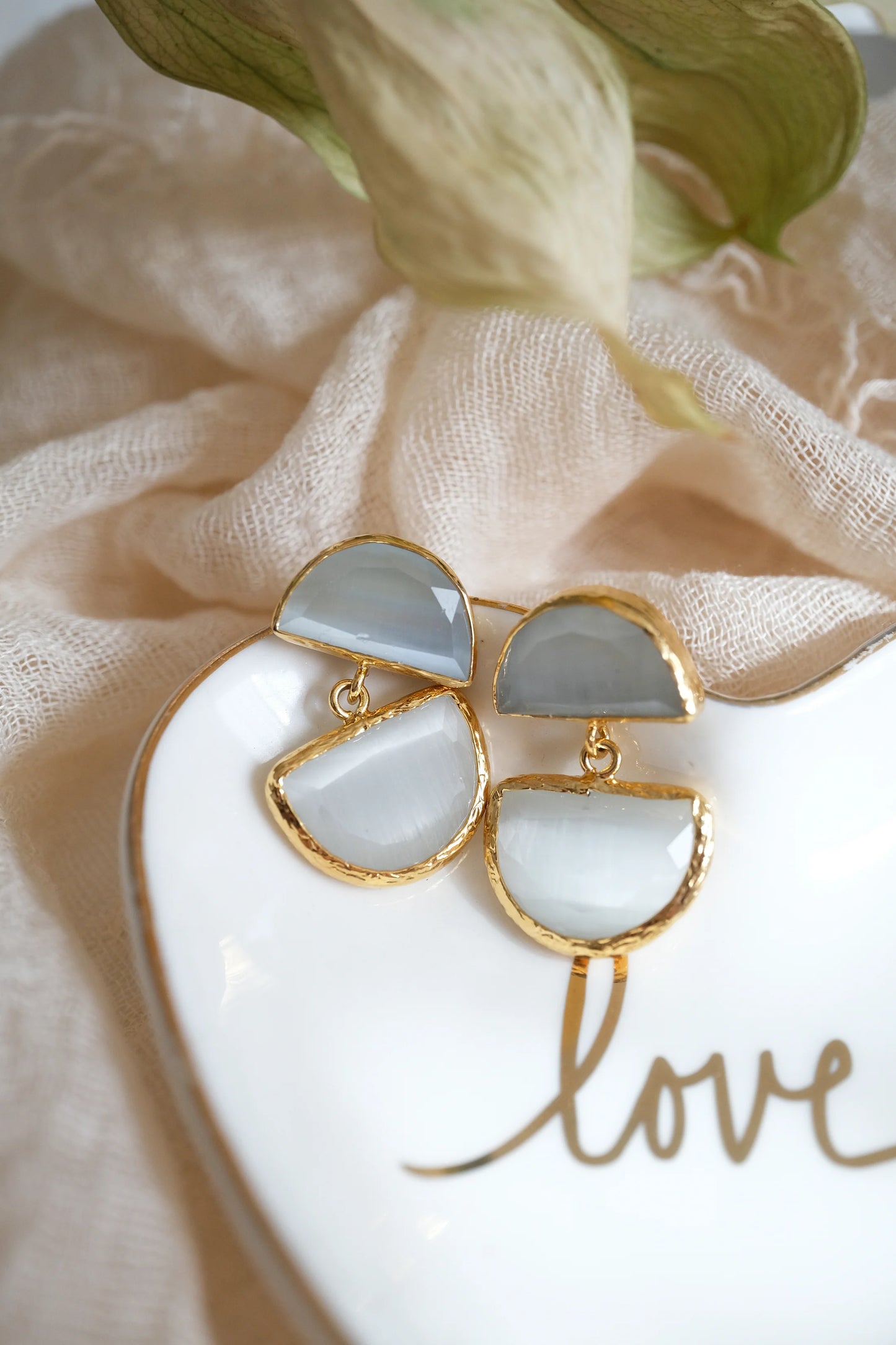 Gold and white earrings on a heart-shaped dish with 'love' text, against a soft fabric background.