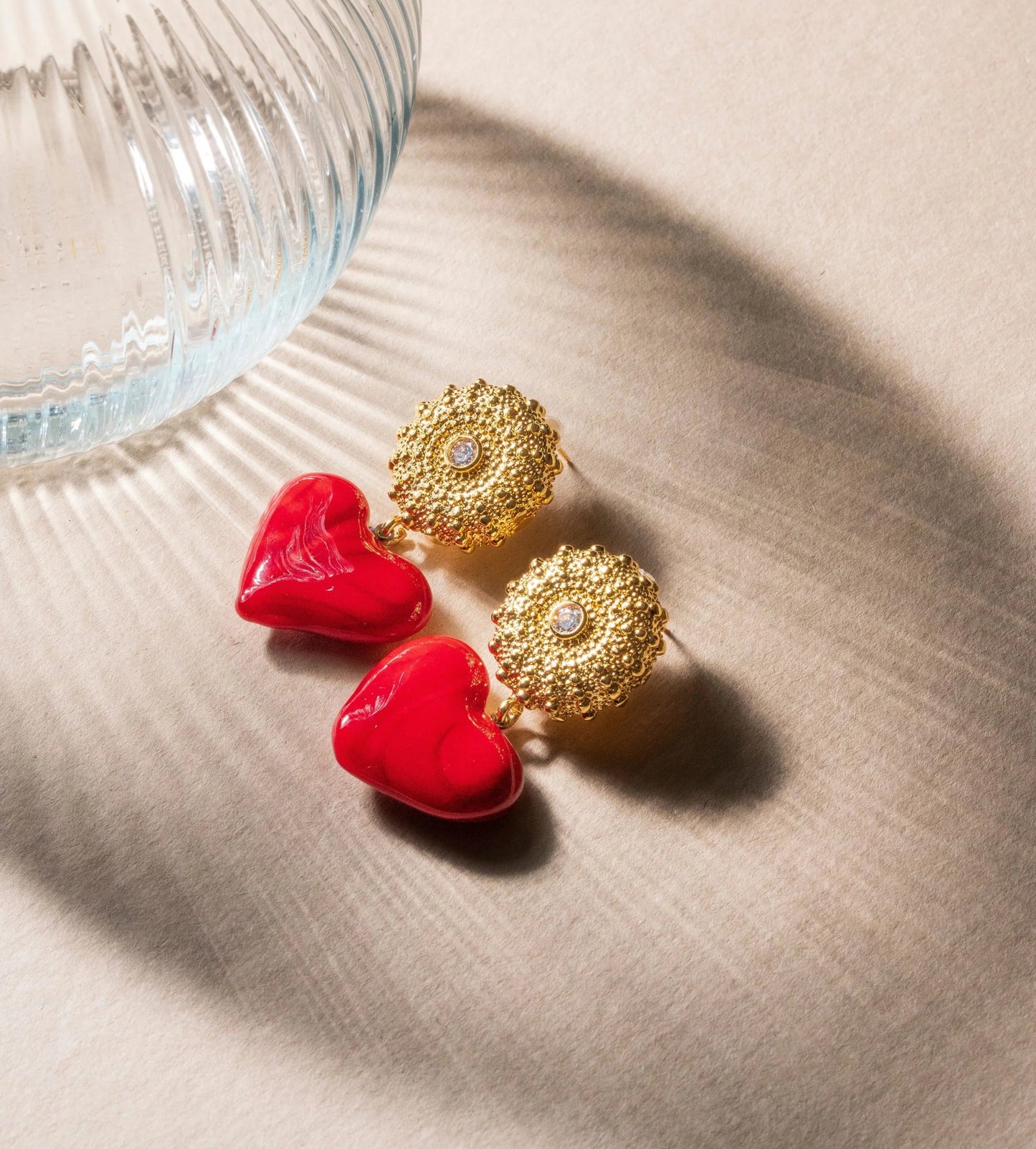 Gold earrings with red heart-shaped pendants on a beige surface.